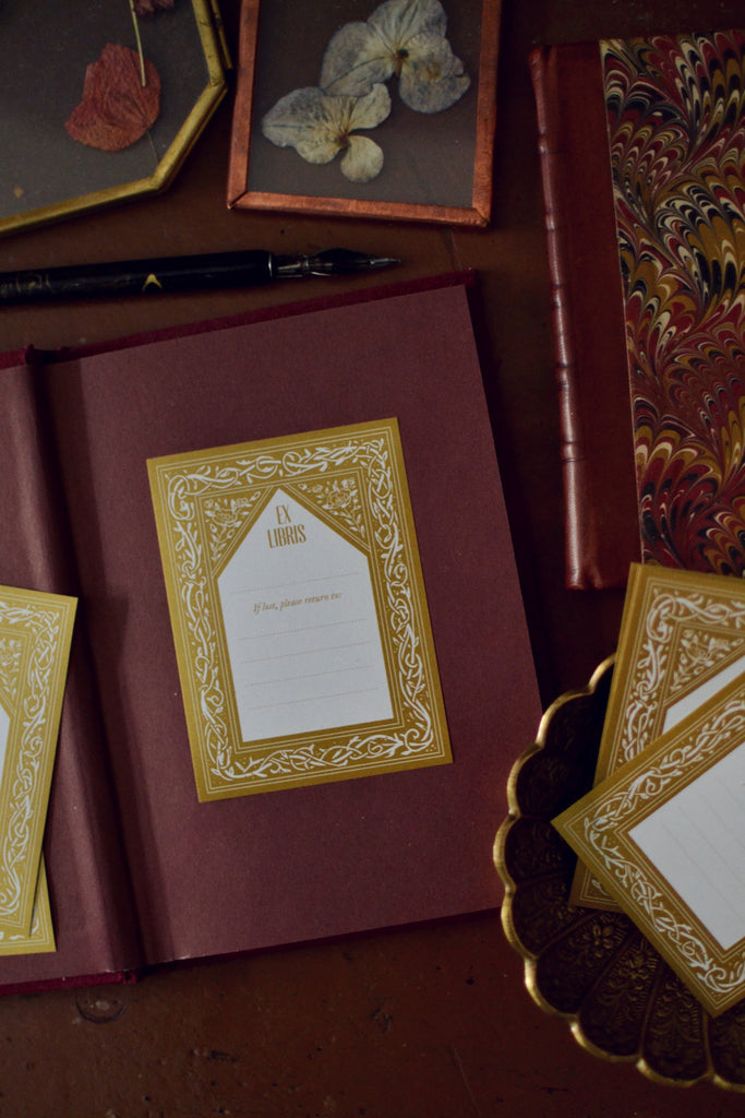 vintage bookplate printed in gold placed on burgundy book and vintage accessories surrounding it and pressed flowers