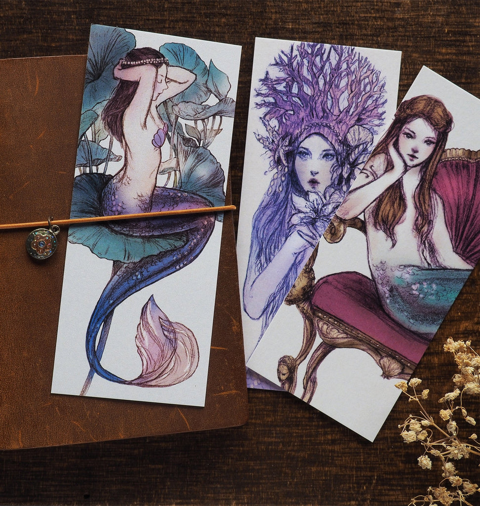 Illustrated mermaid bookmark tucked into an elastic book band flatlay on a desk